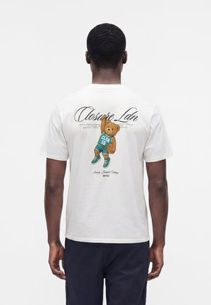 Άνδρας που φοράει λευκό T-shirt με γραφικό ενός αρκουδάκι σε μπασκετικό φανελάκι που κρατάει μπάλα μπάσκετ στην πλάτη.