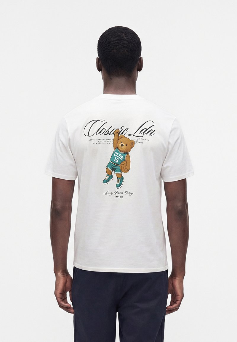 Άνδρας που φοράει λευκό T-shirt με γραφικό ενός αρκουδάκι σε μπασκετικό φανελάκι που κρατάει μπάλα μπάσκετ στην πλάτη.