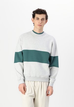 Gray sweatshirt with a teal horizontal stripe. Features a round neckline and ribbed cuffs. Soft texture, relaxed fit. Paired with light-colored trousers.
