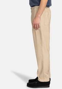 Beige cotton pants with an elastic waistband, side pockets, and a straight leg design. Worn with black moccasin-style shoes.