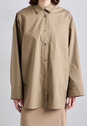 Osoba ubrana w za dużą beżową koszulę zapinaną na guziki z kieszenią na piersi i długimi rękawami oraz beżową spódnicę na jasnym tle.