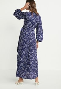 Długa granatowa sukienka w kwiaty z balonowymi rękawami i przewiązką, wyróżniająca się misternym białym wzorem kwiatowym na niebieskim tle.