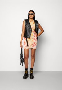 Jaqueta sem mangas de couro preto sobre um vestido mini com padrão tie-dye em tons de laranja e rosa, combinada com botas plataforma pretas e uma pequena bolsa preta.