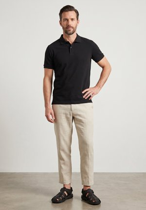 Mand stående med hånden på hoften iført sort poloshirt, beige bukser og sorte sandaler mod en ensfarvet lys baggrund.