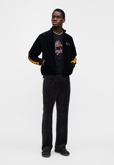 Μαύρο βελούδινο μπουφάν με φερμουάρ και πορτοκαλί λεπτομέρειες, σετ με αντίστοιχο μαύρο βελούδινο παντελόνι με πλαϊνές ρίγες και μαύρο γραφικό μπλουζάκι.