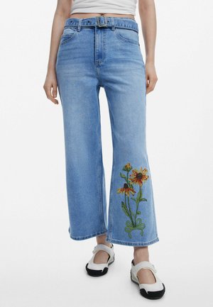 Jean bleu clair à jambes larges avec une ceinture en tissu et des fleurs jaunes et orange brodées près de l’ourlet droit, porté avec des chaussures blanches à brides.