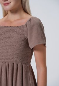 Bruine smockjurk met korte puffmouwen. Textuurstof met een geplooide rok en vierkante halslijn. Subtiele stikdetails op het lijfje.