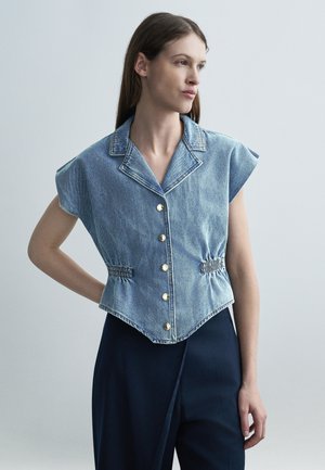 Femeie purtând un vestă din denim fără mâneci, cu nasturi și părți elastice, și pantaloni închiși la culoare, stând cu o mână pe șold, uitându-se pe lateral.