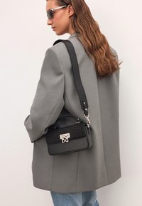 Borsa a tracolla nera con un pannello frontale testurizzato, hardware argentato e una tracolla rimovibile, abbinata a un blazer oversize grigio.