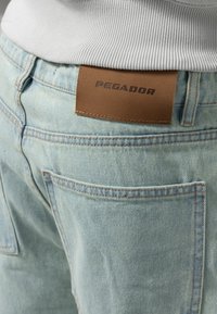 Lyst blå denimjeans med en struktureret overflade, der har en brun læderlap med teksten "PEGADOR" og synlige syninger.
