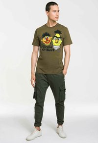 Olive green t-shirt featuring a printed design of two cartoon characters with distinct features. Worn with dark cargo pants and white sneakers.
