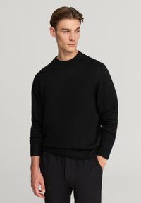 Czarny dzianinowy sweter z okrągłym dekoltem, prążkowanymi mankietami i dolnym obszyciem oraz fakturowanym wykończeniem. Nosi się go na ciemne, casualowe spodnie.