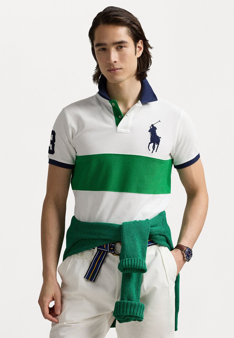 Balts un zaļš svītrains polo krekls ar tumši zilu apkakli, ar zirga logotipu. Zaļa džemperis, kas sasiets ap vidukli, bēšas bikses un pulkstenis.