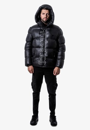 Doudoune noire avec capuche, présentant une finition brillante et du texte en Helvetica blanc. Stylée avec un pantalon cargo noir et des baskets.