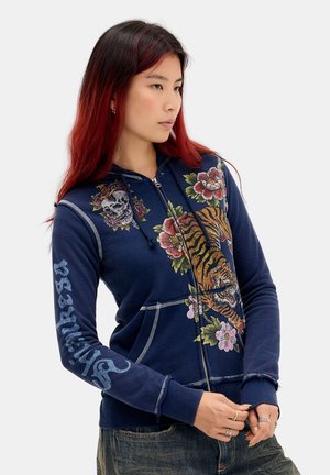 Jeune personne aux cheveux roux portant un sweat à capuche bleu marine avec un motif tigre, floral et crâne, partiellement zippé, les mains ajustant les manches.