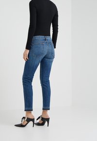 Jeans skinny de mezclilla azul con una cintura de altura media, que cuentan con dos bolsillos traseros y dobladillo deshilachado. Combinados con zapatillas de tacón negras con detalle de lazo.