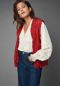 Gilet in maglia rossa con frange sopra una camicia bianca a fiori con dettagli a bottoni, abbinato a jeans in denim scuro a vita alta. Accessori semplici.