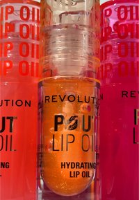 Tre contenitori di lip oil cilindrici: arancione con glitter, rosa e rosso. Il testo nero recita "Pout Lip Oil" e "Hydrating Lip Oil."
