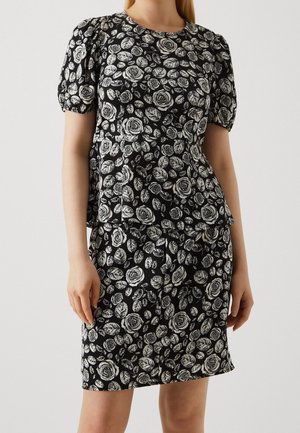 Vrouw draagt een zwarte jurk met witte rozen- en bladpatronen, met korte pofmouwen en een getailleerde taille.