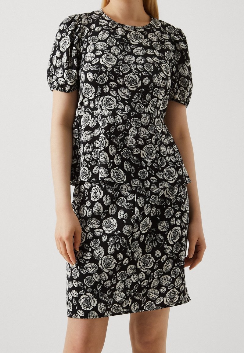 Femme portant une robe noire avec des motifs de roses blanches et de feuilles, avec des manches courtes bouffantes et une taille ajustée.