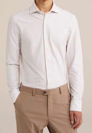 Man die een wit overhemd met lange mouwen draagt met dunne verticale beige strepen en lichtbruine broek met knoop en riemlussen.