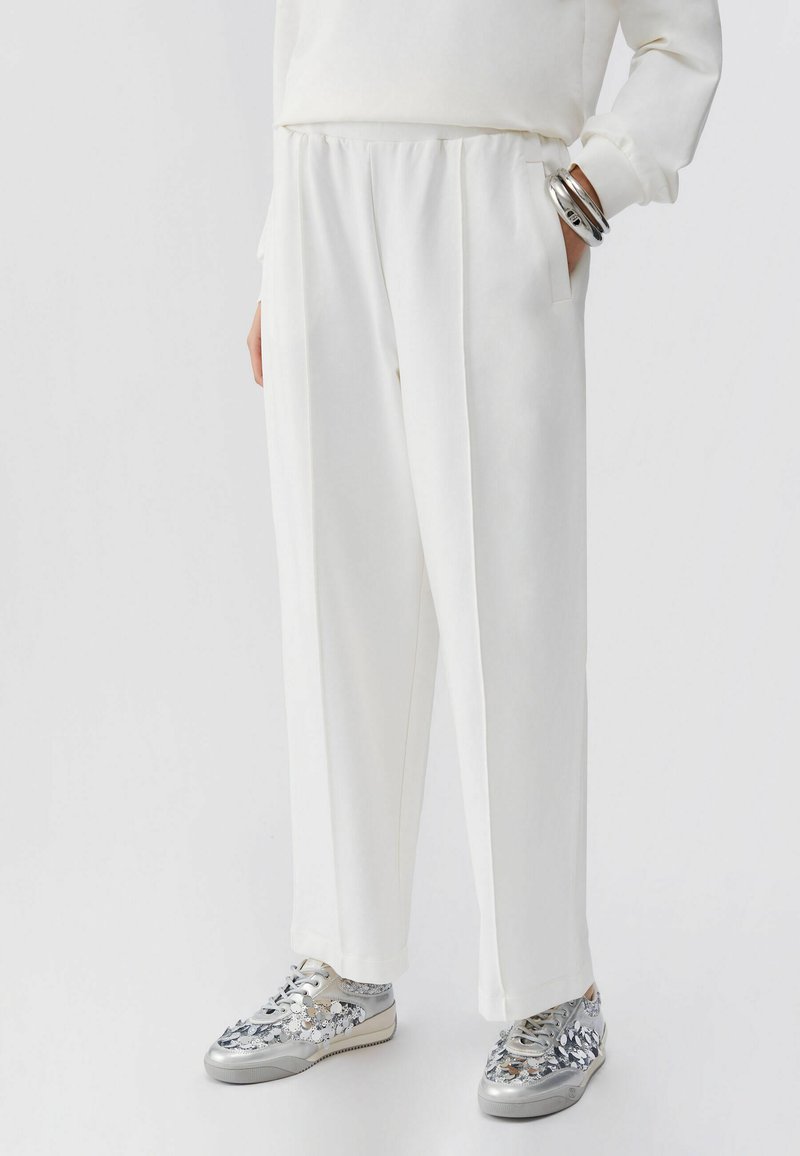 Personne portant un pantalon blanc ample avec poches et des baskets argentées à motif, debout devant un fond blanc uni.