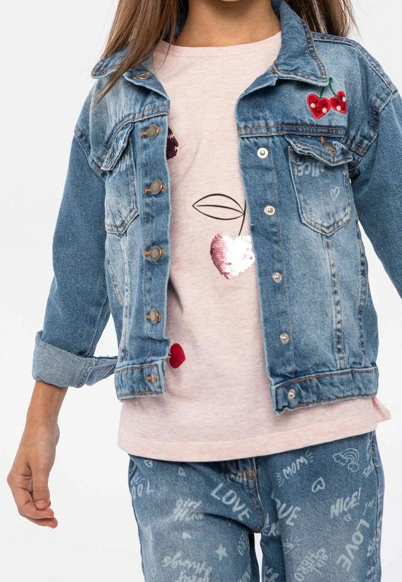 Giacca di jeans con lavaggio chiaro, chiusura a bottoni e maniche arrotolate; abbinata a una maglietta rosa con cuori di paillettes e un design testuale.