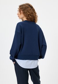 Sudadera de gran tamaño en azul marino con hombros caídos, puños y dobladillo acanalados. Superpuesta sobre una camisa de rayas claras. Textura de tela suave.