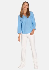Helles blaues Blusenshirt mit V-Ausschnitt, dreiviertelÄrmeln und FaltenDetail, kombiniert mit weißen Hosen und weißen Sneakers.