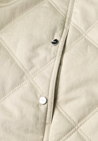 Nærbillede af beige quiltet stof med diagonal syning og to sølvfarvede trykknapper langs en søm.