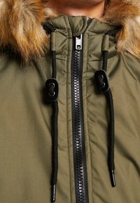 Parka vert olive avec une capuche en fausse fourrure, équipée d'une fermeture éclair noire et de cordons de serrage, présentant une texture lisse et un design matelassé.