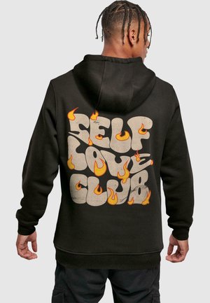 Black hoodie featuring "SELF LOVE CLUB" text in a textured cream with flame accents in orange and yellow. Soft material, relaxed fit.