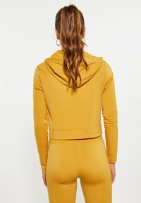 Felpa corta color giallo senape, con cappuccio e maniche lunghe. Tessuto morbido con una silhouette aderente, abbinata a leggings coordinati.