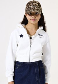 Sudadera corta blanca con cremallera y emblema de estrella negra, combinada con una falda de mezclilla. El modelo lleva una gorra de estampado de leopardo y collares de oro en capas.