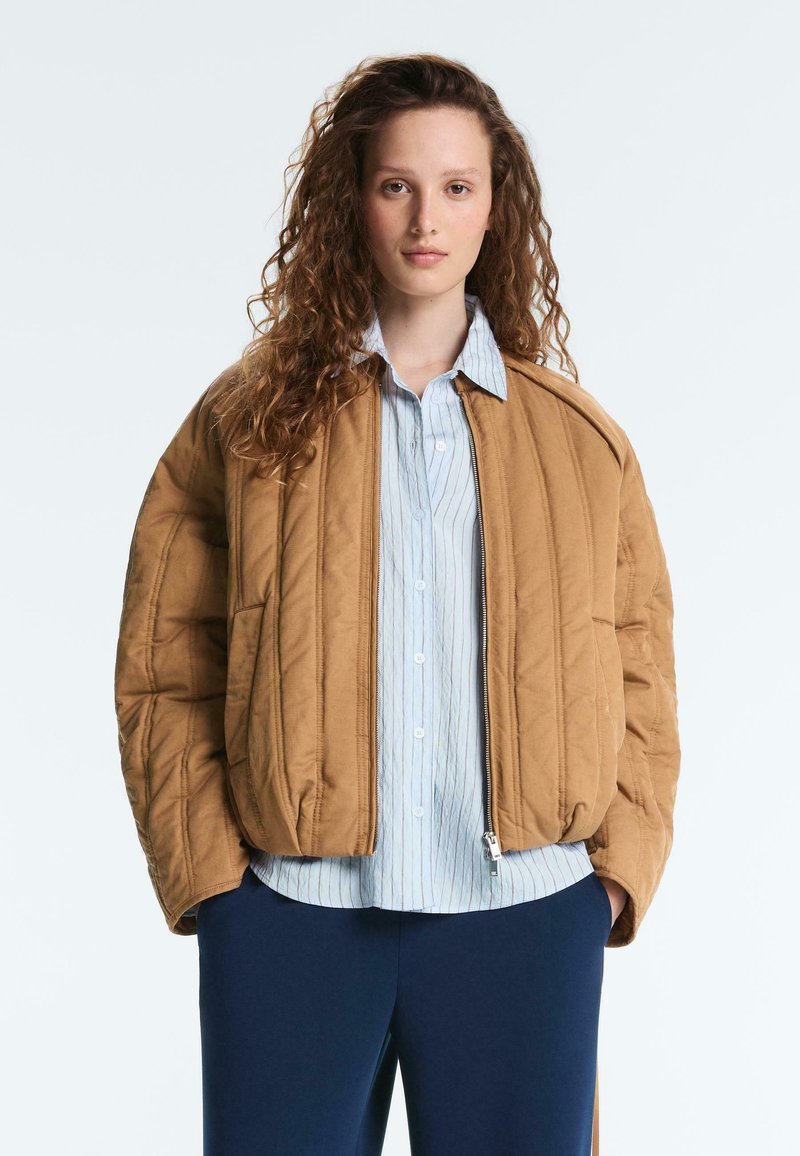 Jeune femme aux cheveux bouclés portant une veste matelassée beige sur une chemise à rayures bleues boutonnée et un pantalon bleu foncé, debout les mains dans les poches.