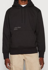 Svart Calvin Klein Jeans-hoodie med framficka, dragsko och beige cargobyxor som bärs av en stående person.