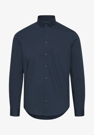 Mężczyzna w dopasowanej, długorękawowej, ciemnoniebieskiej koszuli na guziki i jasnoszarych dżinsach, stojący na tle jednolitego tła.