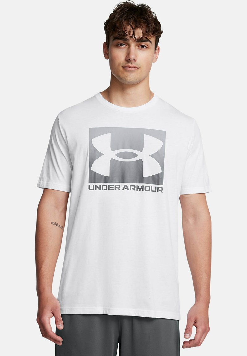 Camiseta blanca de algodón con un gran logo gris de Under Armour centrado en la parte frontal. Mangas cortas, cuello redondo y corte relajado.