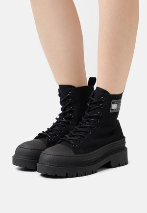 Jambes portant des bottes montantes en toile noires avec des semelles épaisses et crantées et des laçages sur un fond blanc.