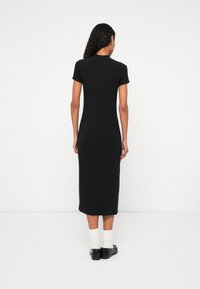 Femme aux cheveux bouclés portant une robe midi noire à manches courtes, des chaussettes blanches et des chaussures noires, debout face à un mur blanc.