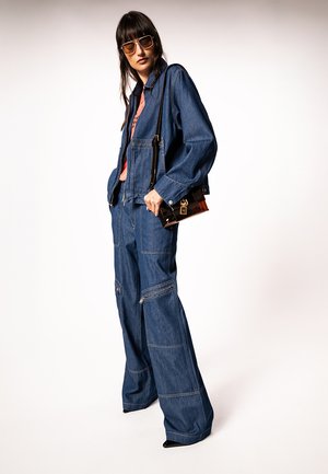 Kobieta ubrana w oversize'ową ciemną kurtkę dżinsową i spodnie o szerokich nogawkach, niosąca małą brązową torebkę i nosząca okulary przeciwsłoneczne.