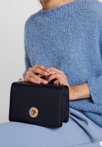 Námořnická kožená kabelka se zlatými detaily a kruhovým logem. Kompaktní obdélníkový tvar s texturovaným povrchem a odnímatelným popruhem.