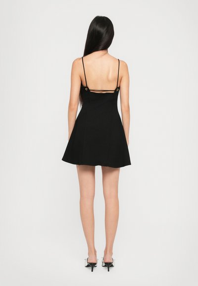 Robe skater noire avec des bretelles fines, un corsage ajusté et une jupe évasée. Détail de découpe notable dans le dos, maintenu par des accents en métal doré.