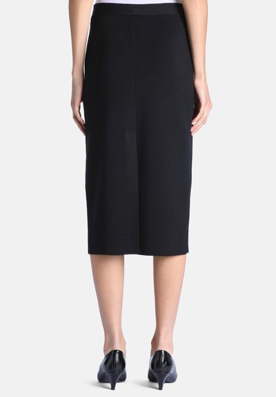 Femme portant une jupe crayon noire longueur genou et des chaussures noires à talons bas, debout les mains détendues le long du corps, vue de dos.
