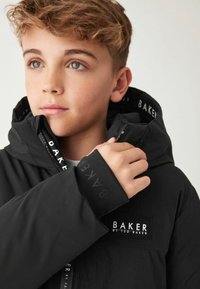 Veste noire avec un patch logo en évidence. Comprend une capuche, une fermeture éclair et un tissu texturé. La doublure affiche le nom de la marque de manière répétée.