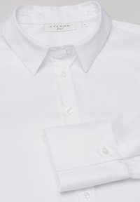 Camicia bianca aderente realizzata in tessuto morbido, con colletto classico, nonché una fila di bottoni e polsini con bottoni. Etichetta del brand visibile all'collo.