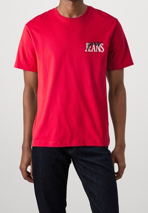 Mand iført en lys rød kortærmet T-shirt med hvidt og sort "JEANS" logo på venstre bryst kombineret med mørkeblå jeans.