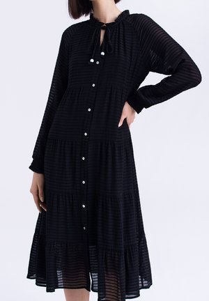 Robe noire à volants en tissu texturé, avec un devant boutonné, des manches longues et un col à nouer, dotée de légères rayures horizontales.