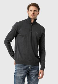 Dunkelgrauer Zip-Kragen-Pullover mit strukturierter Oberfläche, gerippten Bündchen und Saum sowie dezentem Logodetail auf der Brust. Getragen über blauen Jeans.