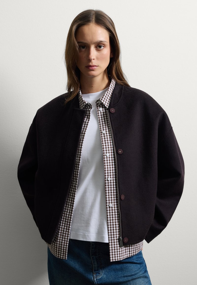 Veste en polaire noire avec col rond, fermeture à boutons, portée avec une chemise à carreaux et un t-shirt blanc en dessous, stylisée avec un jean en denim.